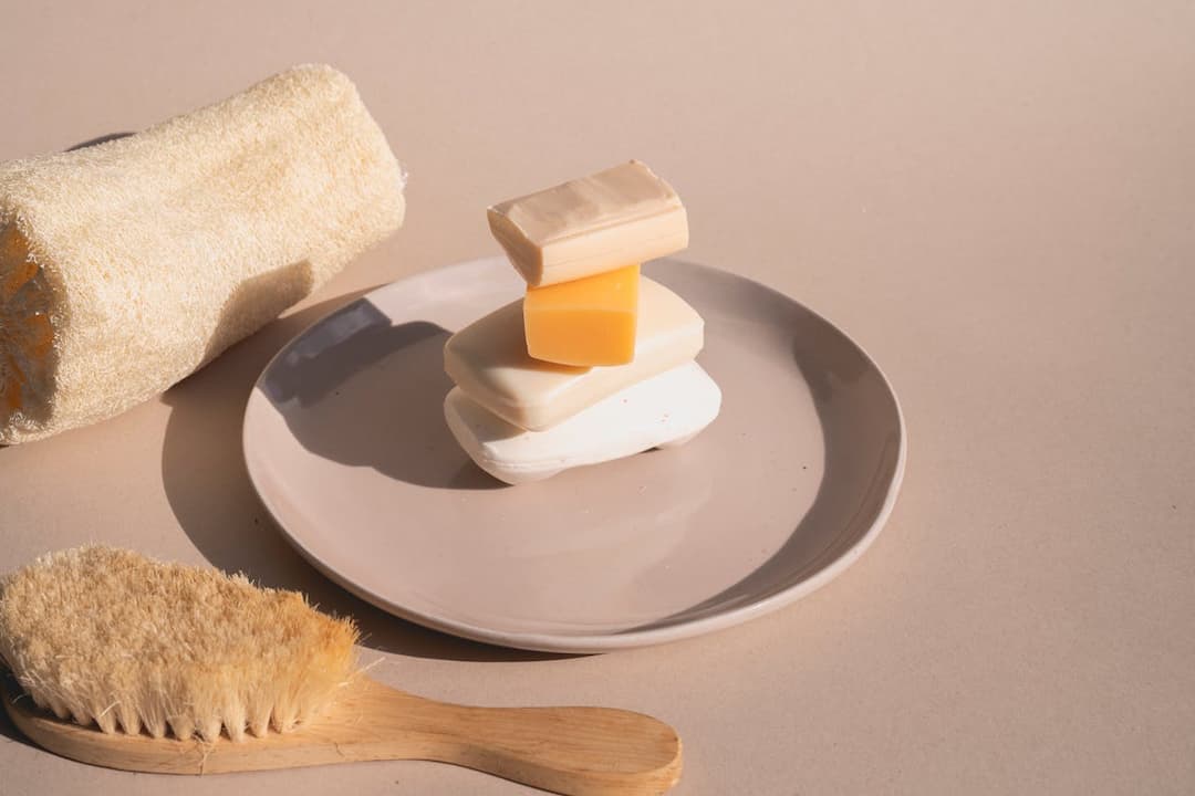 Stack of soap bars on a ceramic plate, with a natural loofah sponge and wooden bath brush on a beige surface