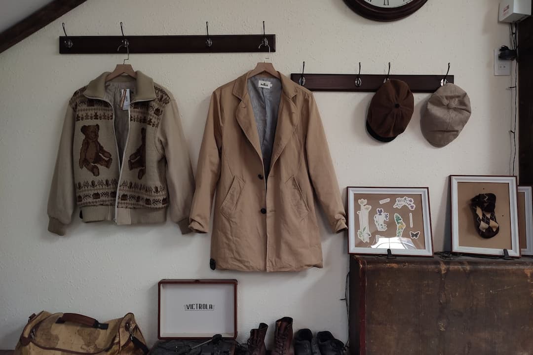 A cozy vintage corner with a teddy bear sweater, tan trench coat, and two flat caps hanging on hooks above a collection of framed art, shoes, a record player, and a worn travel bag