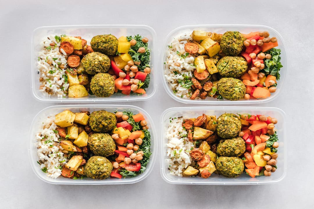 Four plastic meal prep containers filled with rice, roasted vegetables, chickpeas, chopped bell peppers, kale, and three green falafel balls each