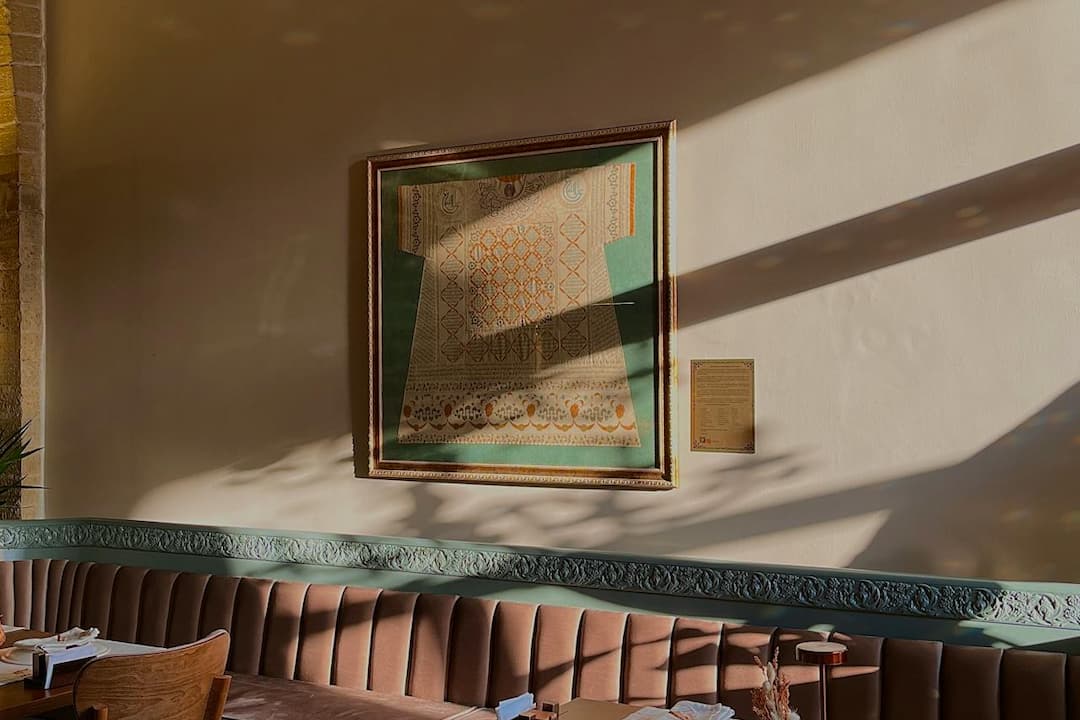 Framed embroidered textile in the shape of a robe, mounted on a taupe wall with green accents, above a tufted bench in a softly lit room