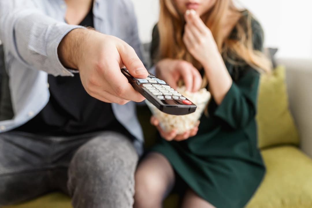 man pointing TV remote toward camera, woman beside him eating popcorn, both sitting on couch, casual clothing, blurred background