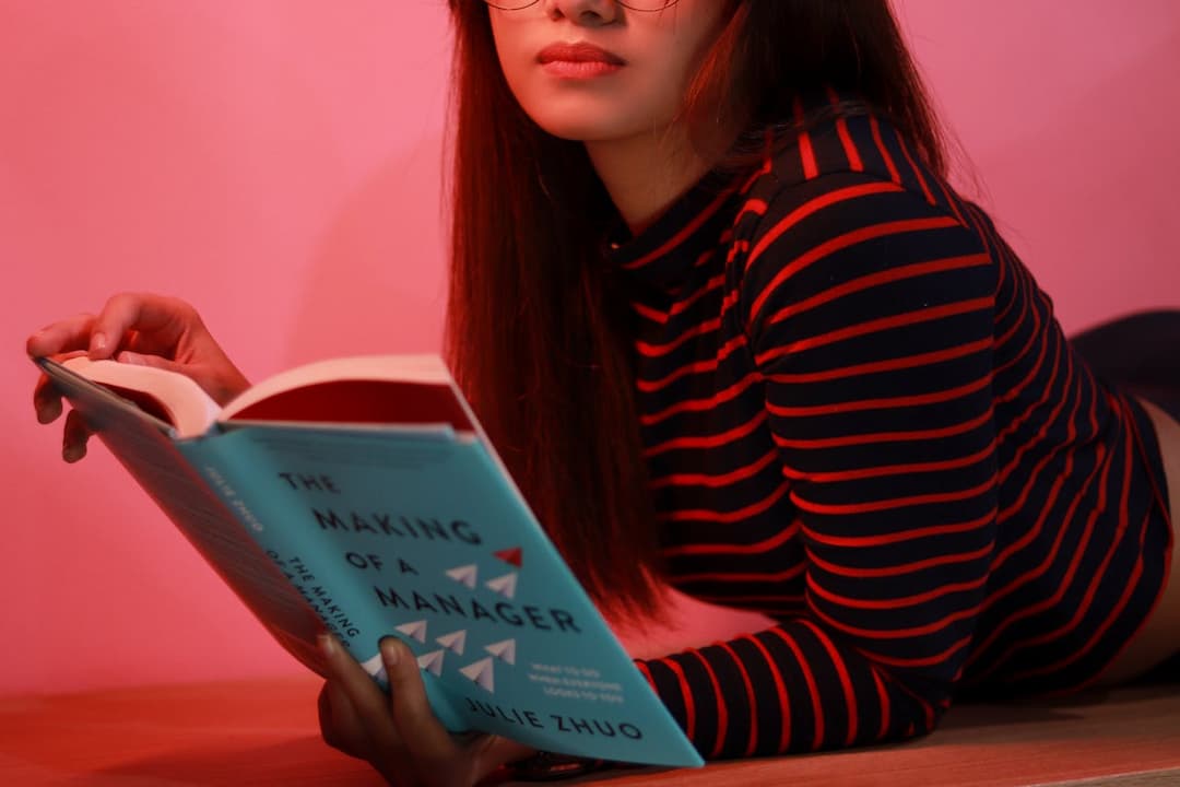 Woman wearing glasses and a striped shirt reading The Making of a Manager by Julie Zhuo under pink lighting
