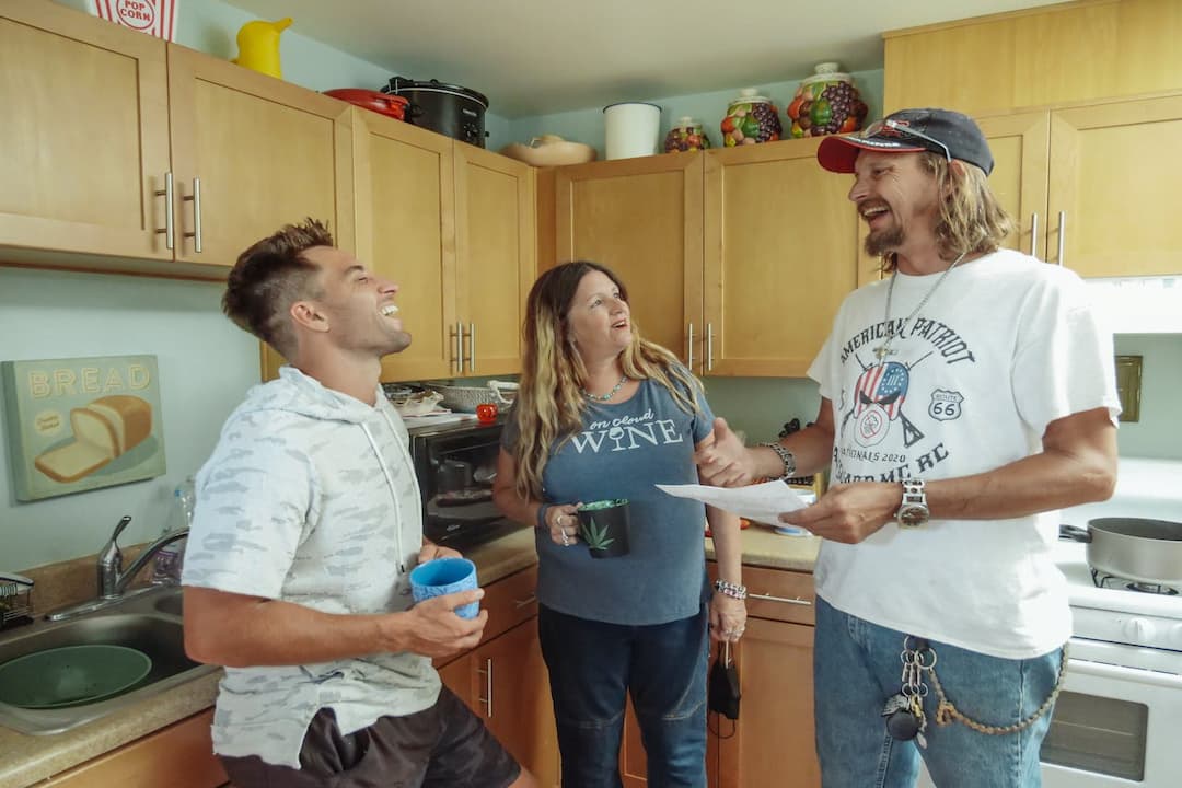 three adults chatting and laughing in a home kitchen, man in white hoodie holding a blue mug, woman in blue shirt with a black mug, man in cap holding papers, wooden cabinets, kitchen decor items on top