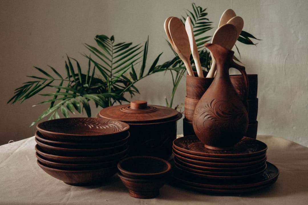 set of carved wooden kitchenware, stacked bowls and plates, lidded pot, decorative pitcher, wooden spoons in cups, palm leaves in background, cream tablecloth