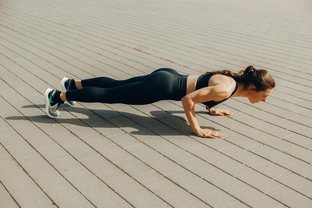 Woman in black athletic wear doing a push-up on a tiled outdoor surface under sunlight