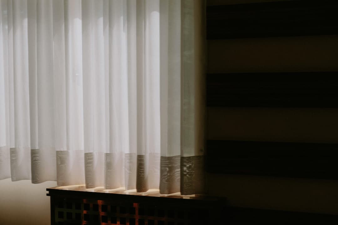 White sheer panel curtains hanging in front of a window, softly lit by natural sunlight, partially covering a wooden vent or cabinet