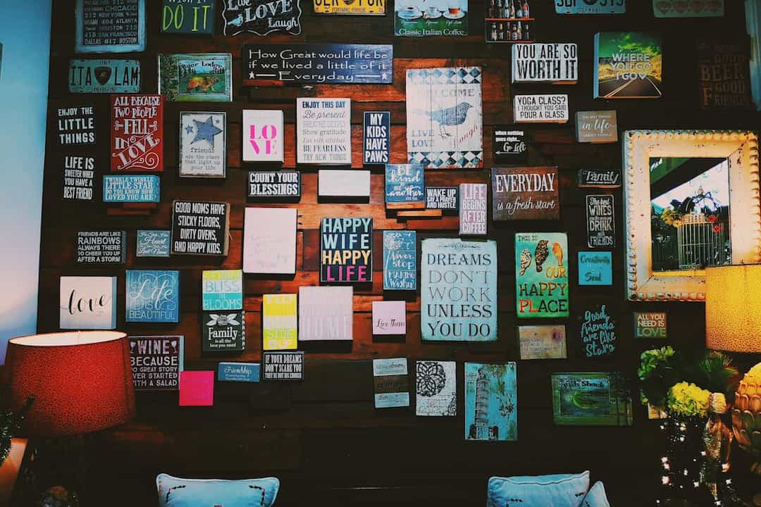 wooden wall decorated with colorful framed quotes, motivational signs, license plates, and artwork, two lamps with red and yellow shades, mirror on right side, white cushions with embroidered designs