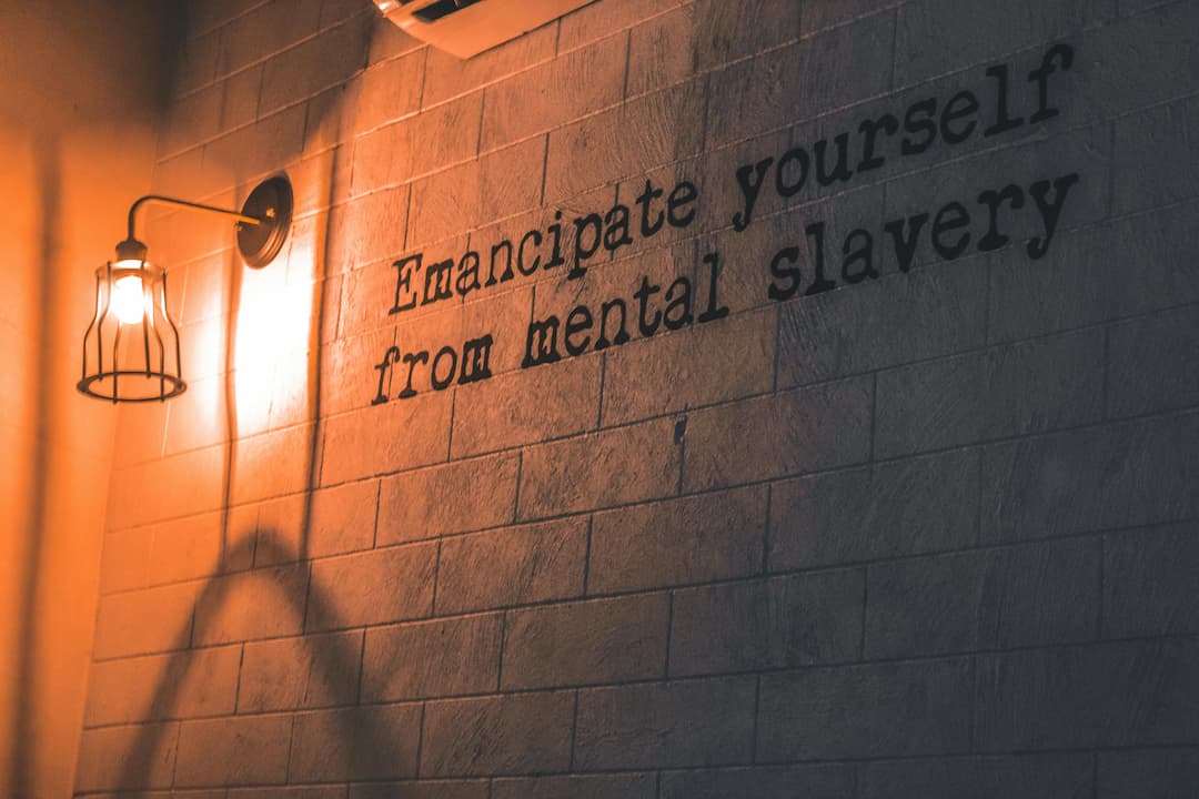 Dimly lit wall with the quote “Emancipate yourself from mental slavery” in black lettering, illuminated by a vintage wall sconce with a caged bulb