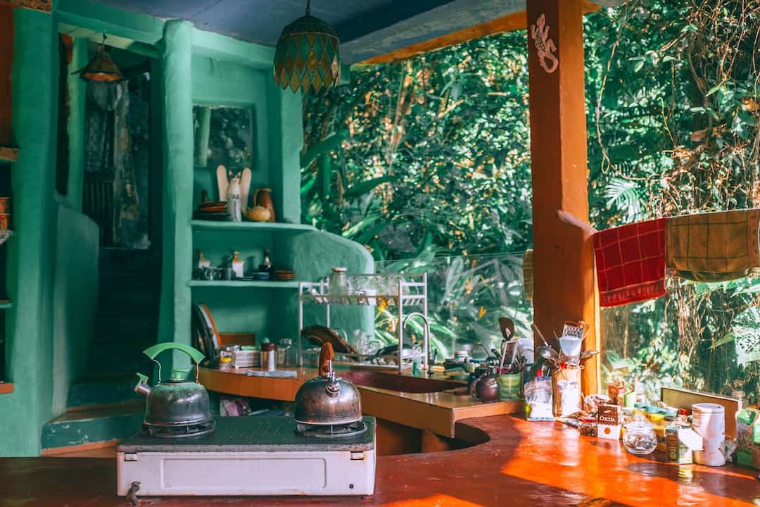 rustic kitchen with green stucco walls, open shelves with utensils and pottery, wooden countertop, two metal kettles on a stove, hanging dish towels, jungle view through glass wall, natural lighting