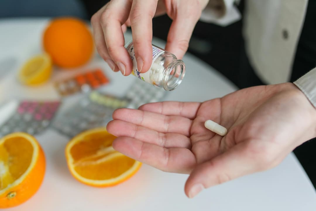 Person holding a white capsule in one hand while pouring it from a small supplement bottle, with sliced oranges and pill blister packs on a white table in the background