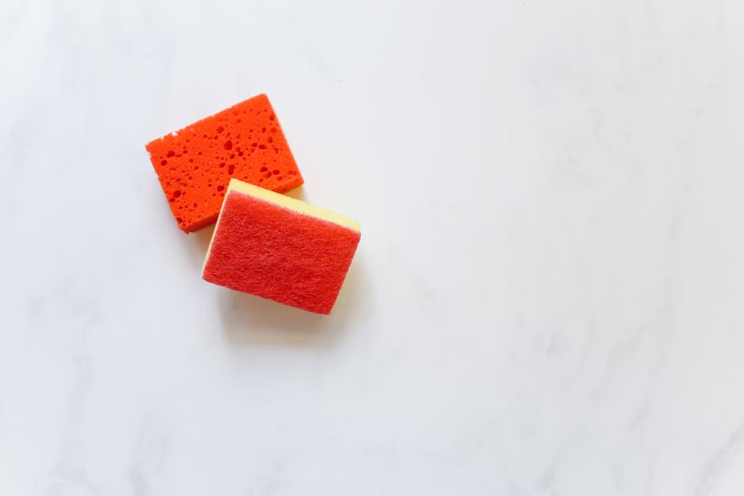 Two rectangular red cleaning sponges on a white marble surface, one with a textured scrubber side and the other with a porous surface