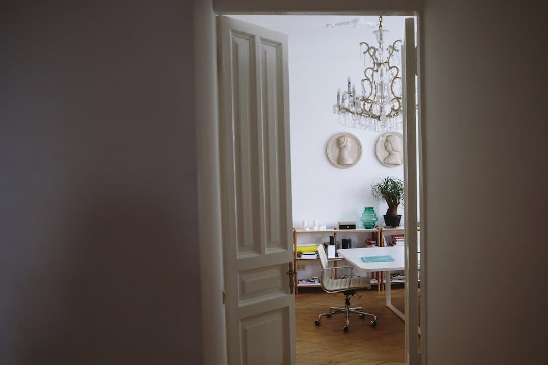 partially open white double doors, view into a bright home office, modern glass chandelier, sculpted wall medallions, white desk with a clear chair on casters, wooden bookshelf with files and decor, potted plant in corner