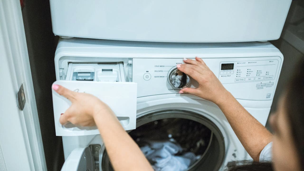 Person operating a front-loading washing machine, one hand adjusting the control dial, the other holding open the detergent drawer, laundry visible inside the machine, scene captured indoors with close-up focus on the hands and washer