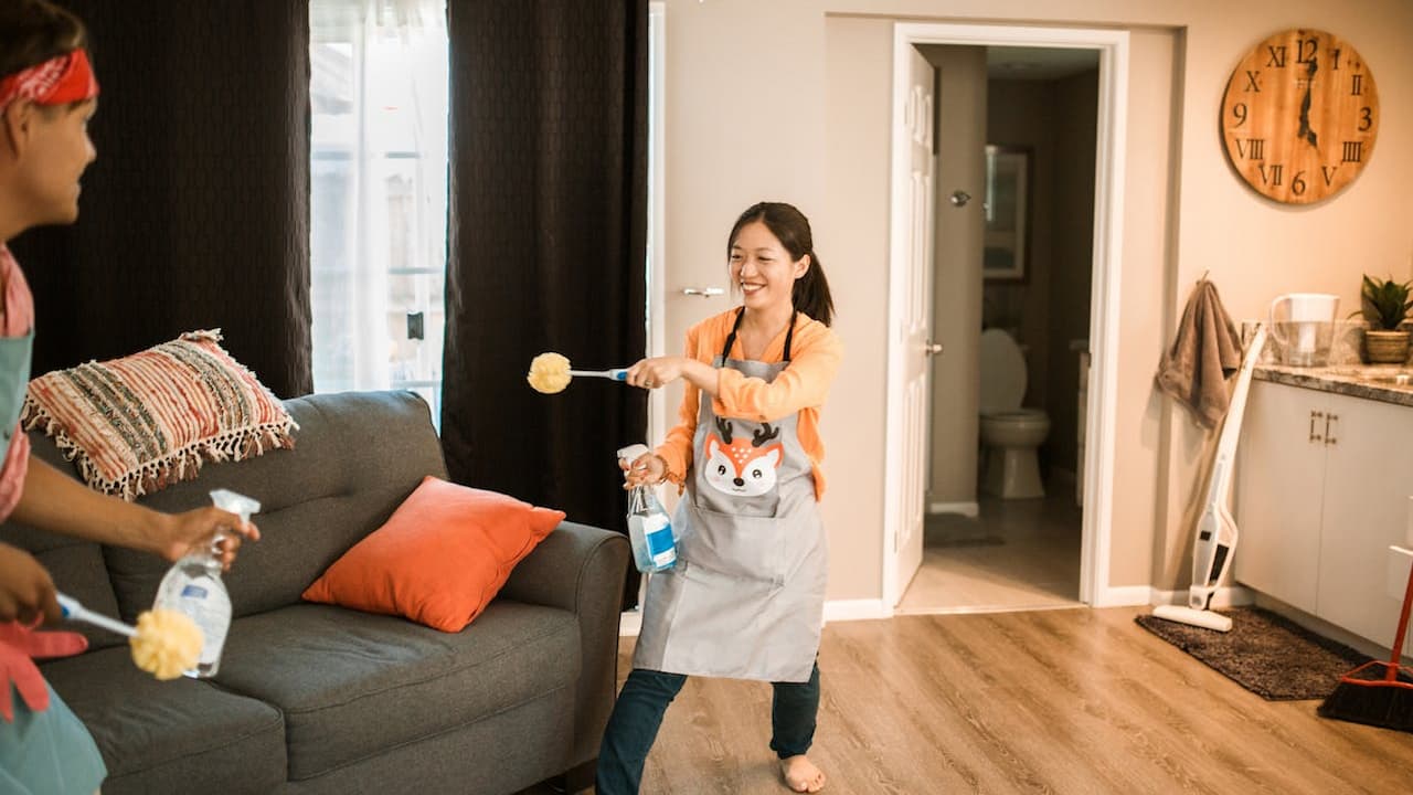 Two people cleaning a living room, both wearing aprons and holding spray bottles and dusters, one playfully pointing a duster at the other, grey couch with orange pillows in the background, open bathroom door visible, wall clock and cleaning tools in the corner, bright natural light coming through the window, casual and cheerful atmosphere