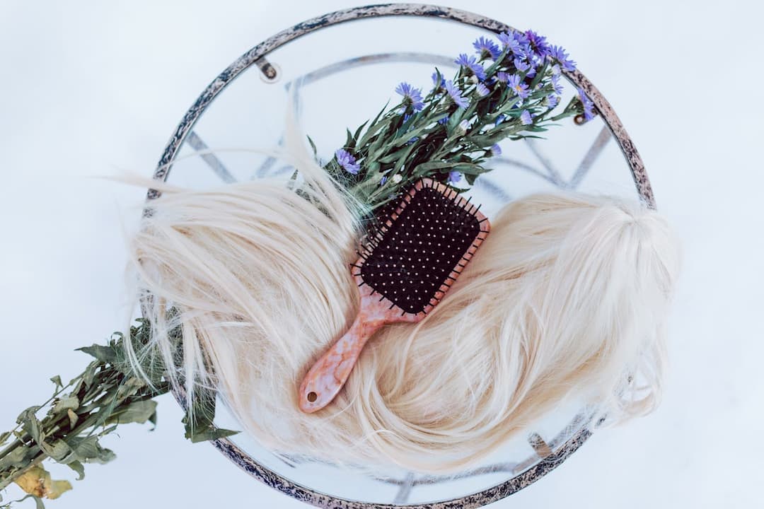  Blonde wig with a pink hairbrush and sprig of purple flowers arranged on a glass table