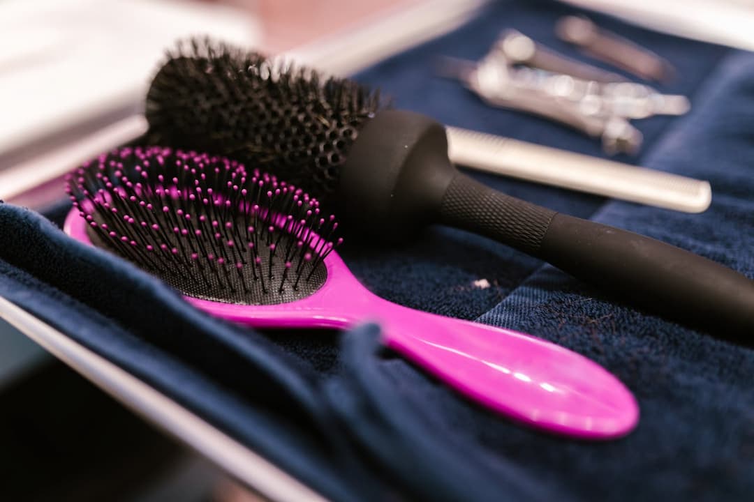 Close-up of a pink detangling brush, a black round hairbrush, and silver salon tools laid out on a dark blue cloth