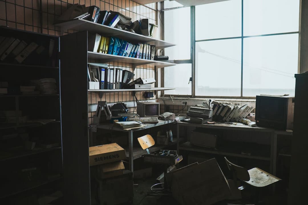 abandoned office room, dusty shelves filled with folders and binders, broken furniture, old computer monitor, vintage TV, cluttered desks, Siemens cardboard boxes, tiled wall, large window with daylight
