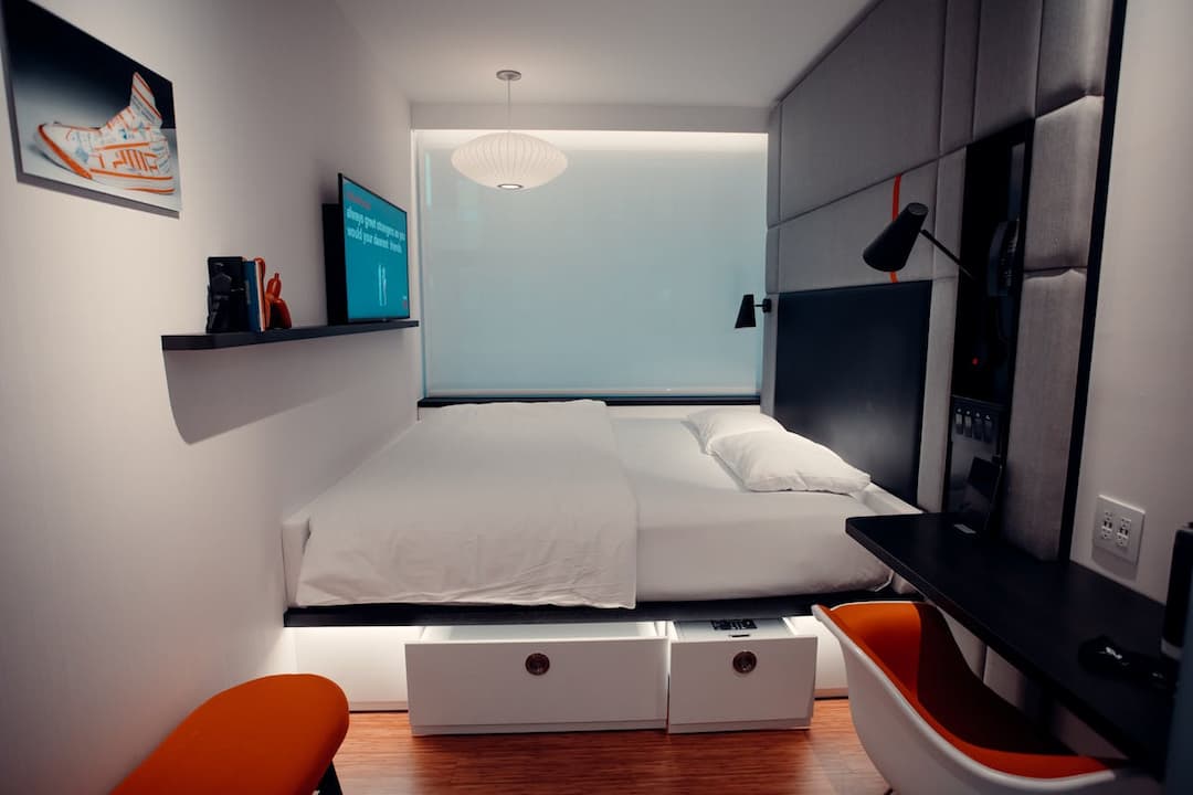 Modern compact bedroom with a double bed featuring white bedding and built-in underbed drawers, a mounted flat-screen TV, floating shelf with books and decor, desk with orange-accented chair, and padded headboard wall