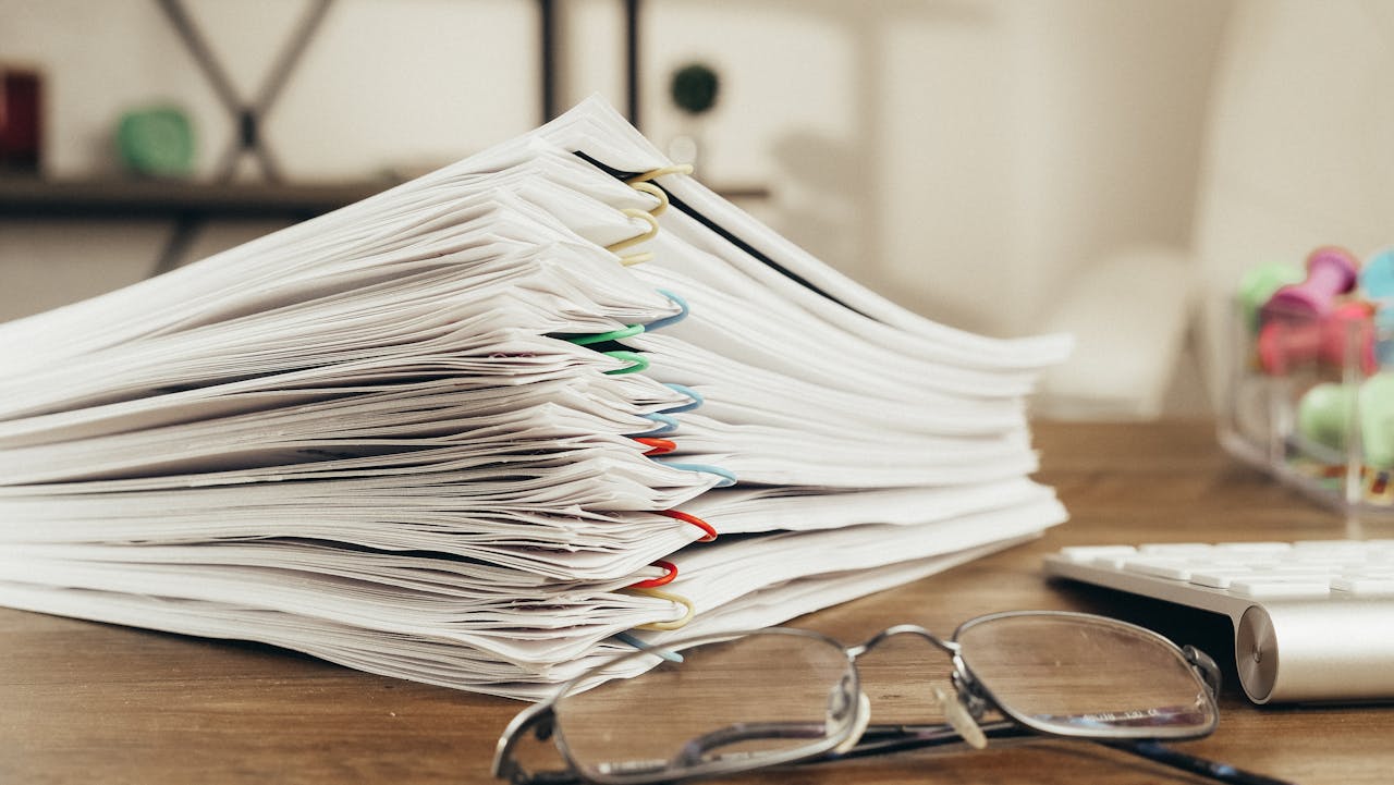 Large stack of documents clipped with colored paper clips on a desk beside reading glasses and a keyboard